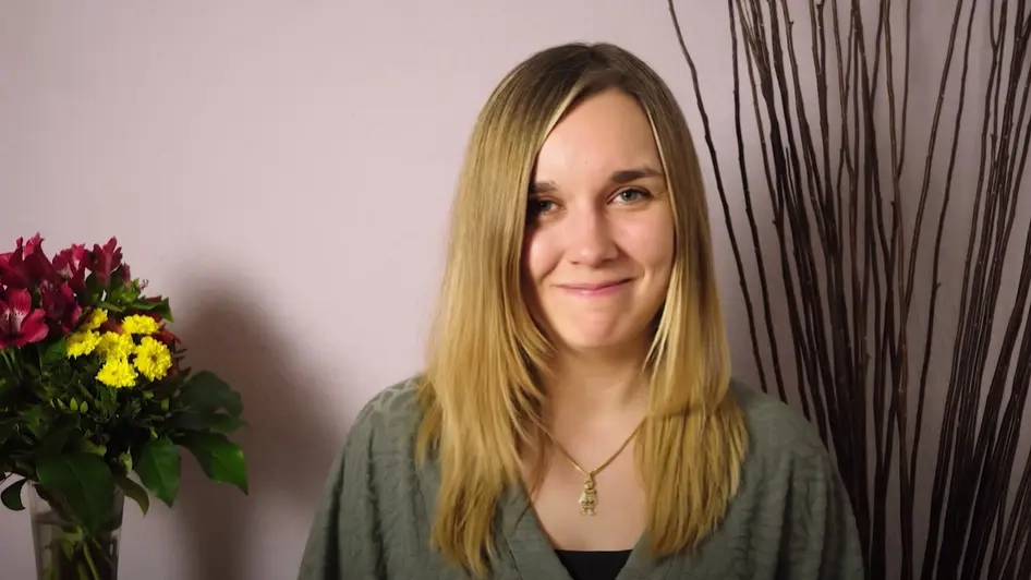 Eine junge Frau mit langen, blonden Haaren lächelt in die Kamera. Neben ihr steht ein Blumenstrauß.