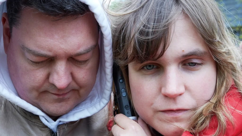Ein Mann und eine Frau stehen dicht beieinander. Zwischen ihren Köpfen hält die Frau ein Tongerät an ihrer beider Ohren.