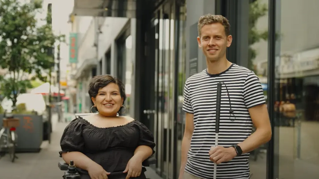 Eine junge Frau im Rollstuhl und ein junger Mann mit Blindenstock stehen in einer Fußgängerzone vor einem Eingang aus Glas.