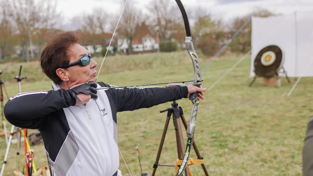 Ein Mann mit Sonnenbrille spannt einen Bogen und zielt auf eine Zielscheibe.