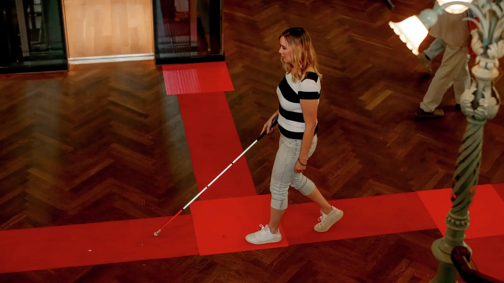 In einem großen Raum geht eine Frau mit einem Blindenlangstock an einer Markierung entlang.