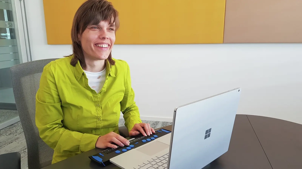 Eine blinde junge Frau sitzt lächelnd vor einem Laptop mit Braillezeile