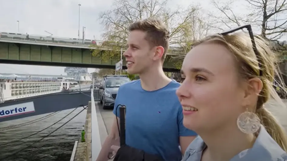 Ein junger Mann und eine junge Frau stehen am Rheinufer. Im Hintergrund sieht man eine Straßenbahn über eine Brücke fahren.