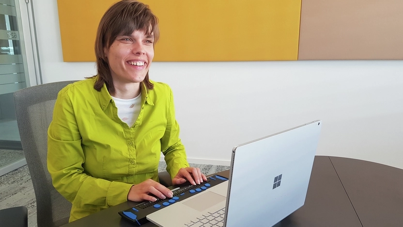 Eine blinde junge Frau sitzt lächelnd vor einem Laptop mit Braillezeile