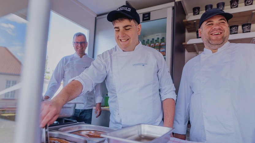 Drei Männer in Kochjacken stehen in einem Foodtruck. Der mittlere bereitet Essen zu.