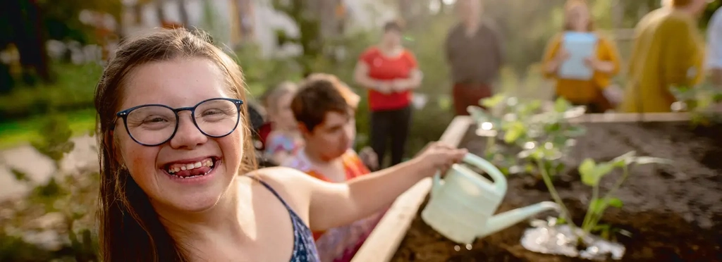 Eine junge Frau mit Down-Syndrom gießt Pflanzen in einem Hochbeet. Sie lacht fröhlich direkt in die Kamera.
