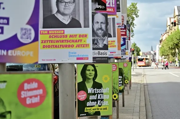 Im rechten Bildrand ist eine Straße zu sehen. Auf dem Bürgersteig auf der linken Bildhälfte sind viele, bunte Wahlplakate an Laternenpfählen angebracht.