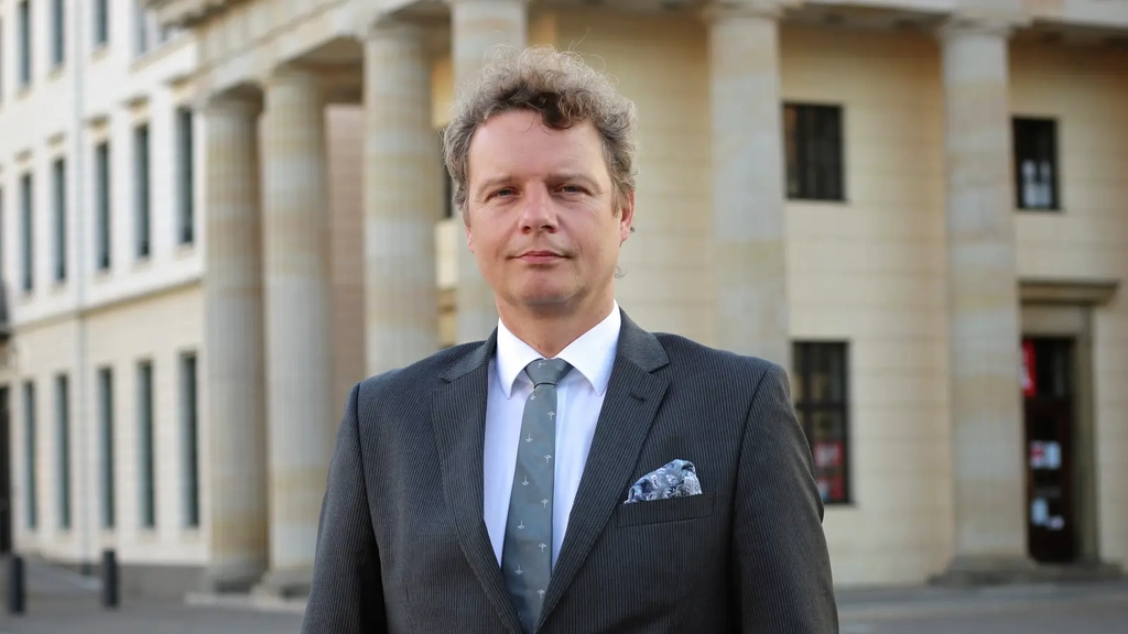 Ein Mann mit lockigen grau-braunen Haaren steht im Anzug vor einem alten Gebäude und schaut in die Kamera.