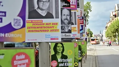 Im rechten Bildrand ist eine Straße zu sehen. Auf dem Bürgersteig auf der linken Bildhälfte sind viele, bunte Wahlplakate an Laternenpfählen angebracht.
