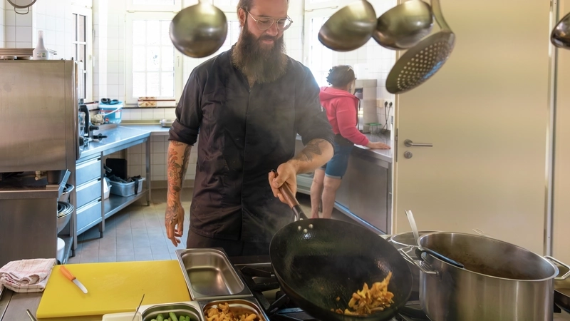 Ein großer Mann mit Brille, Hipster-Bart und tatoowierten Unterarmen schwenkt Gemüse in einer Pfanne über dem Herd einer Restaurantküche.