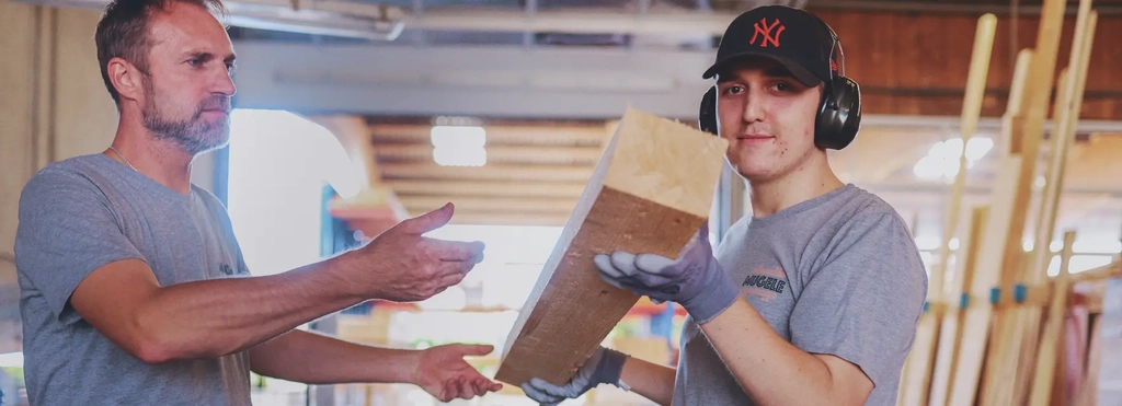 Zwei Männer in einer Holzverarbeitungswerkstatt. Der jüngere der beiden trägt Ohrenschutz und Handschuhe. Er hat einen großen Holzbalken in den Händen. Diesen reicht er in Richtung des anderen Mannes.