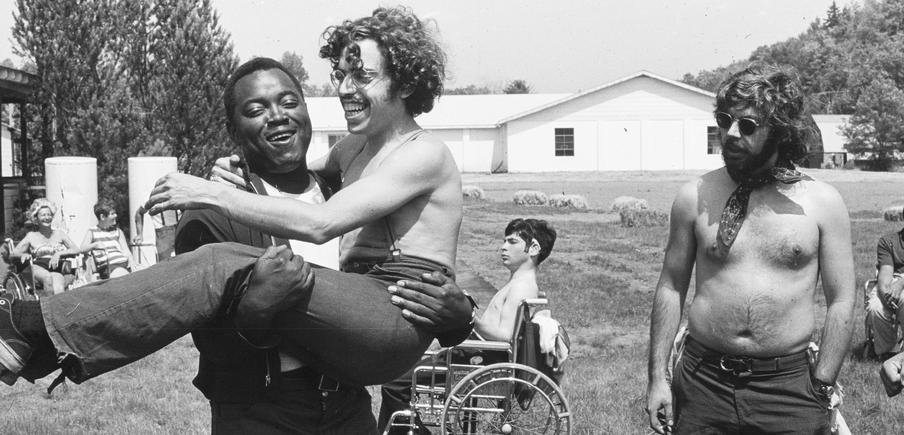 Das historische schwarz-weiße Bild zeigt mehrere Männer und Jugendliche mit und ohne Behinderung draußen. Ein lachender schwarzer Mann trägt einen weißen. 