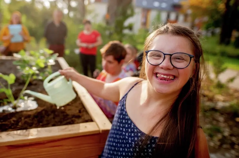 Eine junge Frau mit Down-Syndrom gießt Pflanzen in einem Hochbeet. Sie lacht fröhlich direkt in die Kamera.