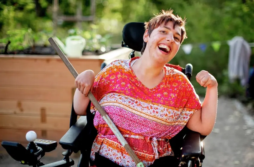 Eine Frau im elektrischen Rollstuhl lacht in die Kamera. In der Hand hält sie eine Harke.