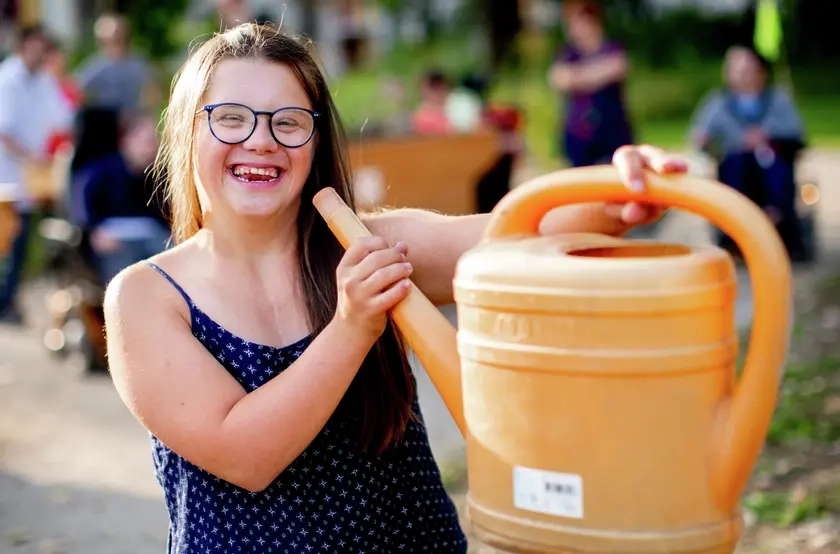 Eine junge Frau mit Down-Syndrom hält eine Gießkanne im Spaß wie ein Megaphon an ihren Mund. Sie lacht fröhlich in die Kamera.