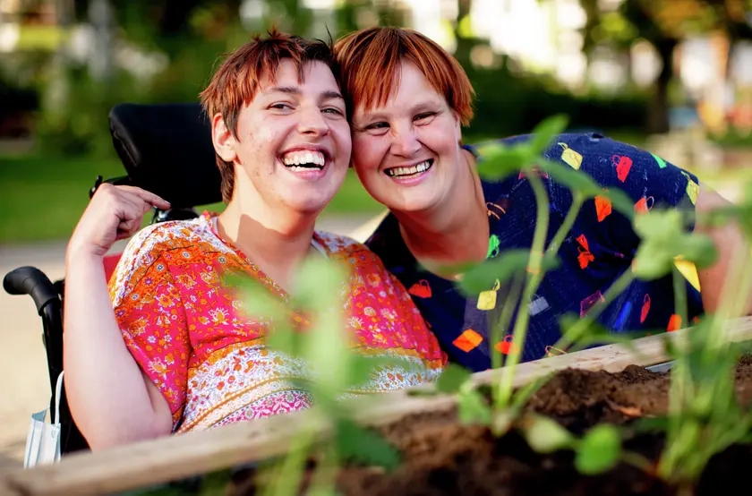 Zwei Frauen in bunt gemusterten Oberteilen sind hinter einem Hochbeet zu sehen. Eine der beiden Frauen sitzt in einem Rollstuhl. Sie stecken die Köpfe zusammen und lachen in die Kamera.