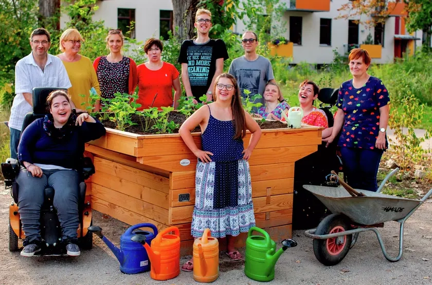 Eine Gruppe von Menschen mit und ohne Behinderung steht um ein Hochbeet, das sie gerade bepflanzt haben. Alle lächeln.