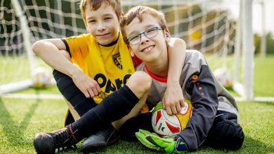 Zwei Jungen in Fußballkleidung sitzen in einem Tor. Sie haben sich freundschaftlich die Arme um die Schultern gelegt und lächeln in die Kamera. Einer von beiden trägt ein Hörgerät und hat verkürzte Beine.
