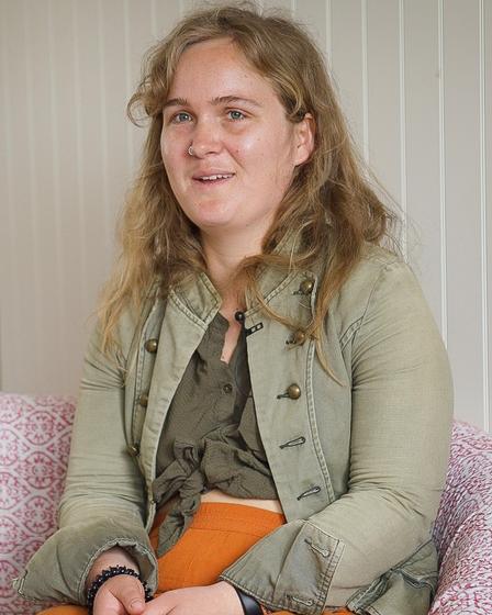 Eine junge Frau mit blonden langen Haaren sitzt in ihrem Wohnzimmer auf dem Sofa. Das Sofa hat einen weißen Bezug mit rosa Muster. Sie trägt eine orangefarbene Sommerhose, ein khakifarbenes Oberteil und eine blassgrüne Jacke.