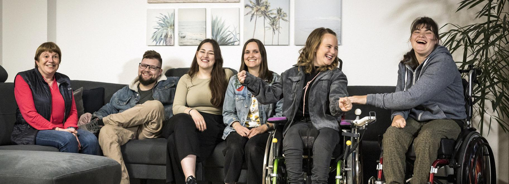 In einem Wohnzimmer sitzen fünf Personen nebeneinander und lachen. Die drei Personen links sitzen auf einem Sofa, die beiden auf der rechten Seite von Rollstühlen, die vor dem Sofa stehen.