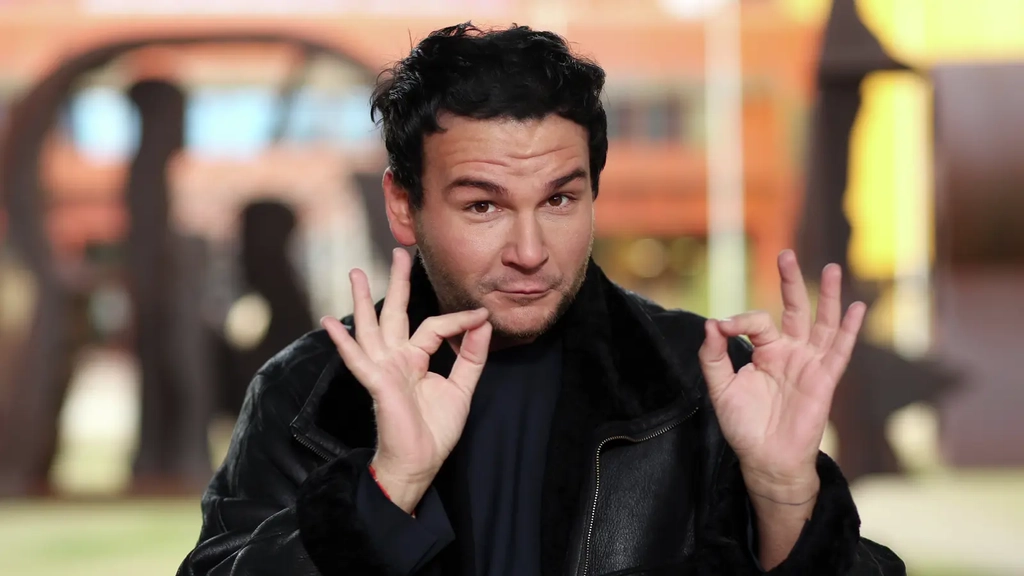 Ein junger Mann mit dunklen Haaren und Lederjacke macht mit seinen Händen eine Gebärde der Deutschen Gebärdensprache.