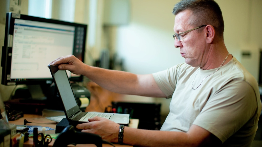 Ein Mann mittleren Alters und mit Brille sitzt an einem Schreibtisch und klappt einen Laptop auf.