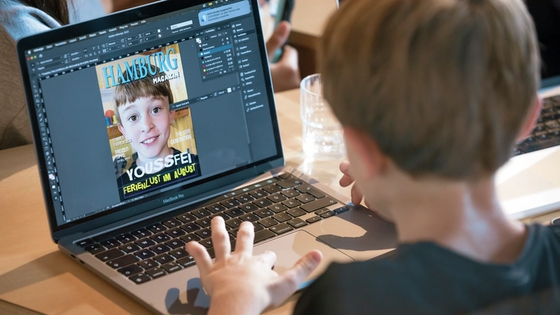 Ein Junge mit dunkelblonden kurzen Haaren und dunklem T-Shirt sitzt vor einem Notebook. Man blickt ihm von hinten über die Schulter. Auf dem Bildschirm ist die Titelseite eines Magazins zu sehen, die der Junge gerade bearbeitet. Das Titelfoto ist das Porträt eines Jungen mit kurzen dunkelblonden Haaren. Vermutlich der Junge, der gerade die Seite bearbeitet.