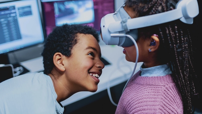 Ein junges Mädchen mit dunkler Haut und langen schwarzen Haaren, die zu dünnen Zöpfen geflochten sind, steht vor einem Tisch mit zwei Computer-Monitoren. Sie trägt eine VR-Brille. Ein etwas älterer Junge, ebenfalls mit dunkler Haut und schwarzen gelockten Haaren beugt sich vor um ihr von unten ins Gesicht zu gucken, als ob er prüfen wollte, ob die Brille richtig sitzt.