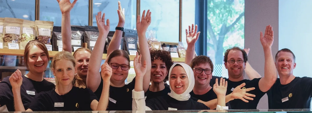 Eine Gruppe von Mitarbeitenden einer Konditorei steht inter der Theke und recken jubelnd die Arme nach oben. Die Personen sind unterschiedlicher ethnischer Herkunft und mit und ohne sichtbare Behinderung.