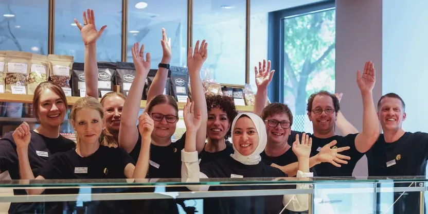 Eine Gruppe von Mitarbeitenden einer Konditorei steht inter der Theke und recken jubelnd die Arme nach oben. Die Personen sind unterschiedlicher ethnischer Herkunft und mit und ohne sichtbare Behinderung.