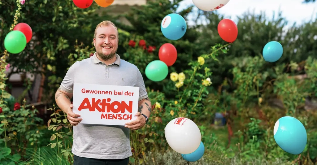 Ein junger Mann steht lachend in einem Garten. Er hält ein Schild in Händen auf dem "Gewonnen bei der Aktion Mensch" steht. Um ihn herum fallen Luftballons zu Boden.