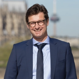 Ein Mann mittleren Alters mit dunkeln Haaren und Brille trägt einen Anzug und schaut in die Kamera.
