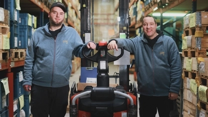 Zwei Männer stehen rechts und links von einem Gabelstapler, der in einer Lagerhalle steht. Die Männer schauen in die Kamera und lächeln.