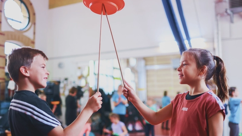 In einer großen Turnhalle, die früher mal eine historische Fabrikhalle gewesen sein könnte, stehen sich ein Junge und ein Mädchen gegenüber. Beide sind etwa acht Jahre alt und tragen Sportkleidung. In einer Hand halten sie jeweils einen Stock, auf dem sie einen Teller balancieren. Beide Stöcke ballanciren denselben Teller. Vermutlich übergeben sie ihn sich gerade.