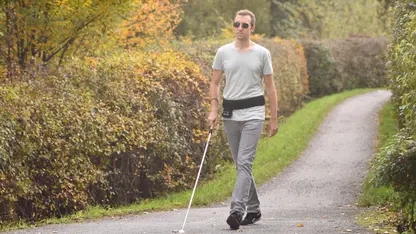 Ein Mann mit Blindenstock geht auf einem schmalen Weg zwischen zwei Hecken. Um seine Hüfte trägt er einen breiten schwarzen Gürtel mit Bedienfeld.