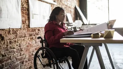 Eine Frau im Rollstuhl sitzt an einem Schreibtisch und schaut sich große Blätter an. An der Wand hinter ihr sind Grundrisse aufgehängt.