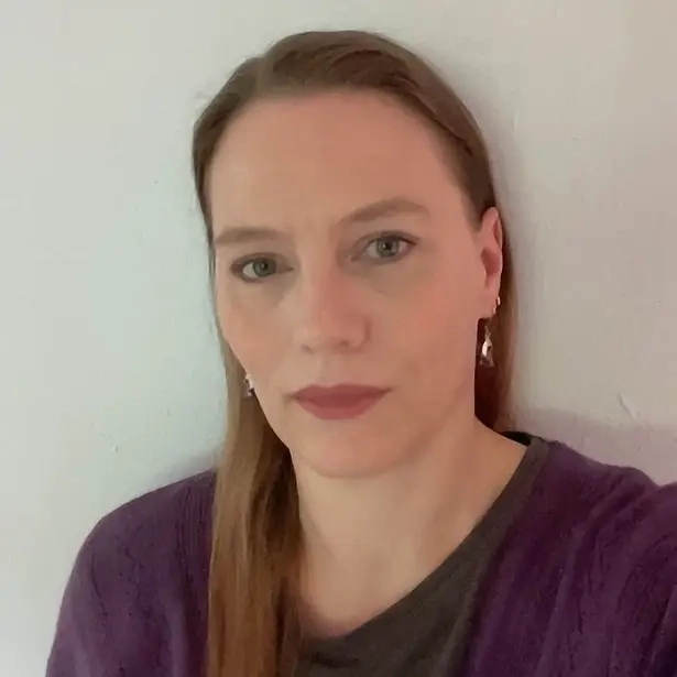 Porträtfoto einer Frau mittleren Alters mit langen glatten dunkelblonden Haaren rot geschminkten Lippen und violetter Strickjacke.