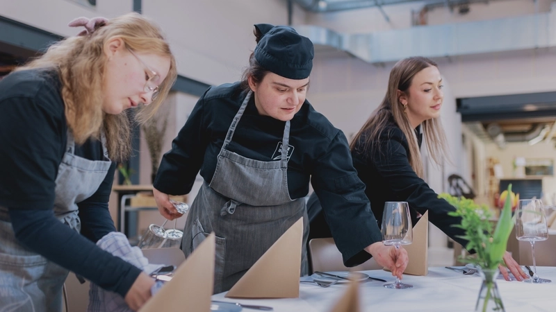 Eine junge Köchin in schwarzer Kochjacke und mit schwarzer Kochmütze stellt ein Weinglas auf eine festlich gedeckte Tafel. Link und rechts von ihr tun zwei junge blonde Frauen aus dem Service dasselbe.