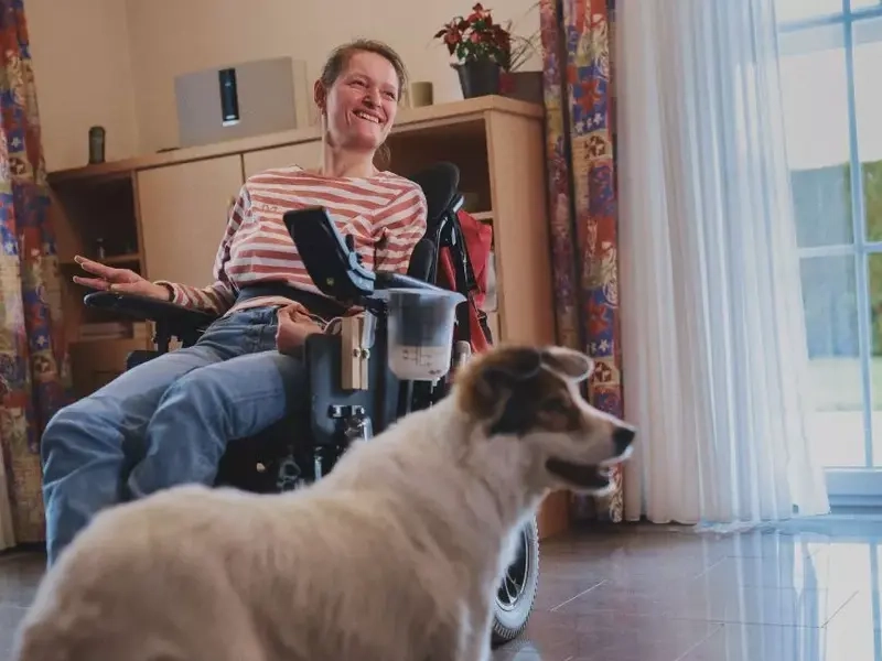 Ein Hund steht in einem Wohnzimmer vor einer Frau im e-Rolli. Er hat die Ohren aufmerksam gespitzt.