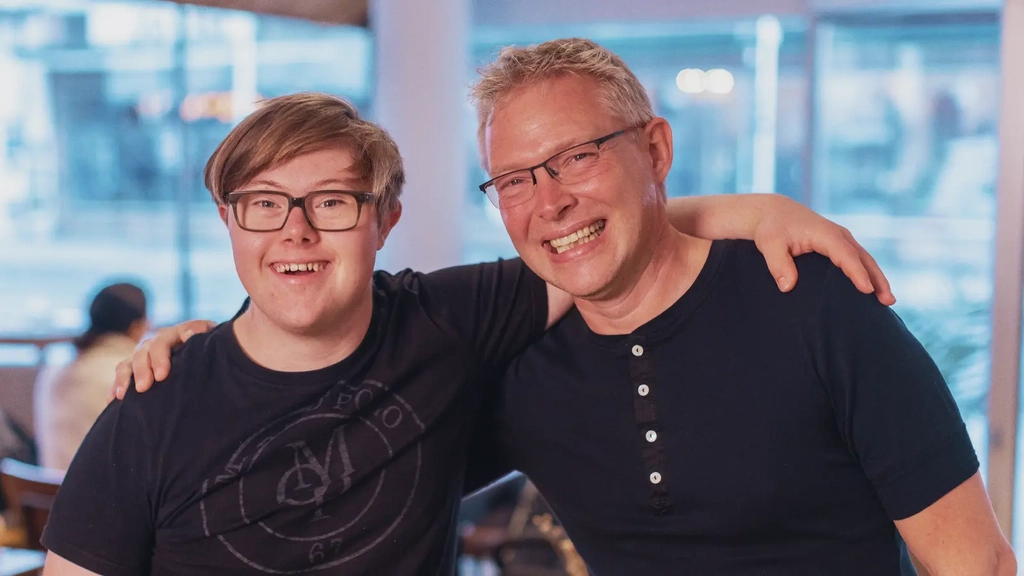 Ein Mann mit Down-Syndrom und ein Mann mit Brille haben einen Arm um die Schultern des anderen gelegt.