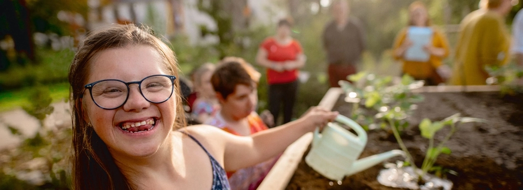 Eine junge Frau mit Down-Syndrom gießt Pflanzen in einem Hochbeet. Sie lacht fröhlich direkt in die Kamera.