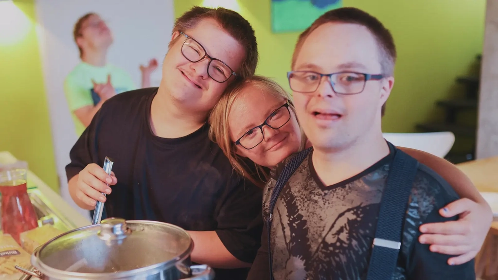 Drei junge Personen mit Down-Syndrom stehen Arm in Arm zusammen in der Küche am Herd und lächeln in die Kamera.