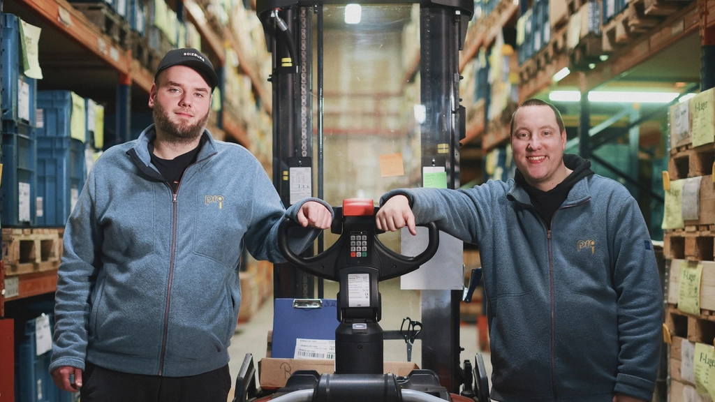 Zwei junge Männer stehen neben einem Gabelstapler in einer Lagerhalle.