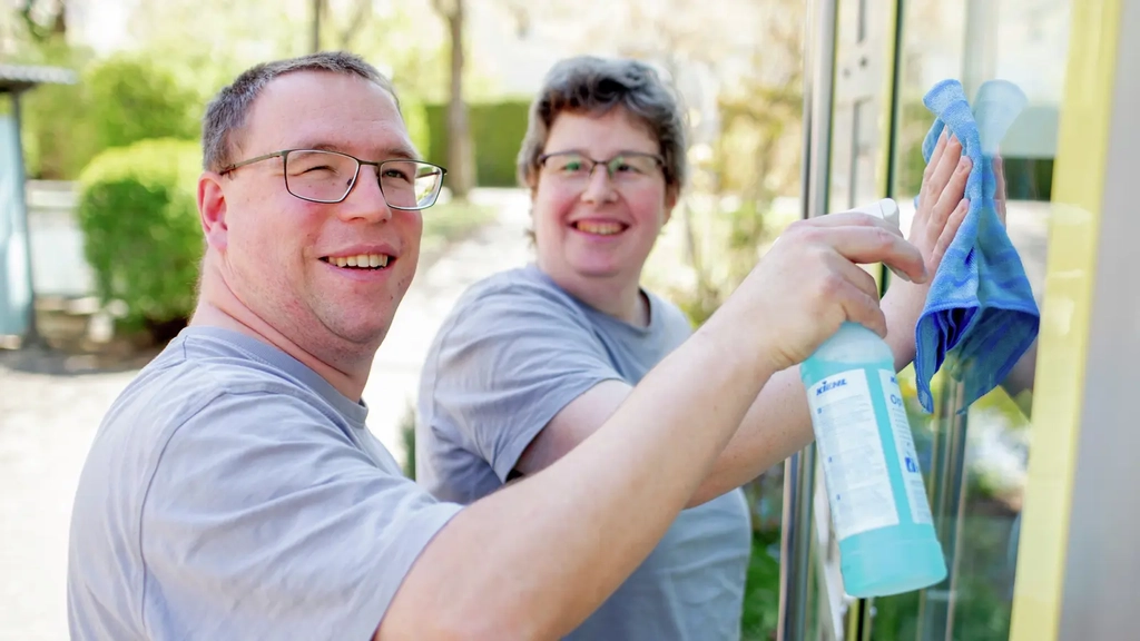 Zwei Personenputzen mit Sprühflasche und Putzlappen eine Fensterscheibe. Beide lachen.