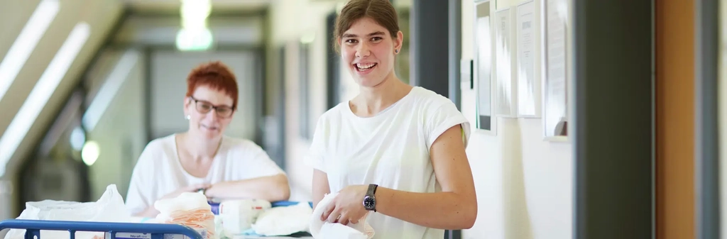 Zwei Frauen stehen in einem Korridor mit einem Wagen voller Verbandsmaterial und ähnlichem Pflege- und Versorgungsmaterial.