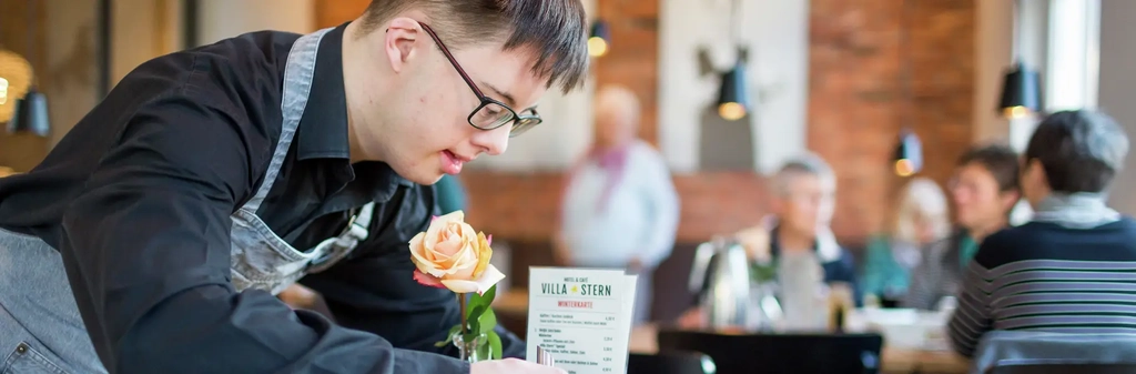 Ein junger Mann mit Downsyndrom steht über einen Cafetisch gebeugt. Er trägt eine Schürze und sortiert Speisekarte und Zuckerstreuer.