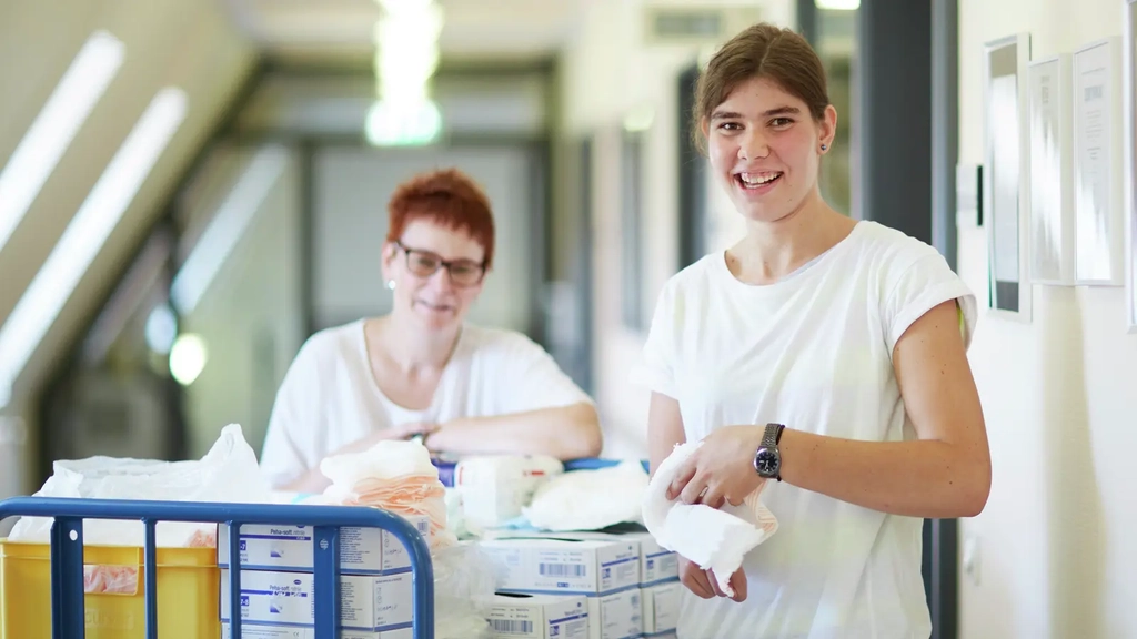Zwei Frauen stehen in einem Korridor mit einem Wagen voller Verbandsmaterial und ähnlichem Pflege- und Versorgungsmaterial.