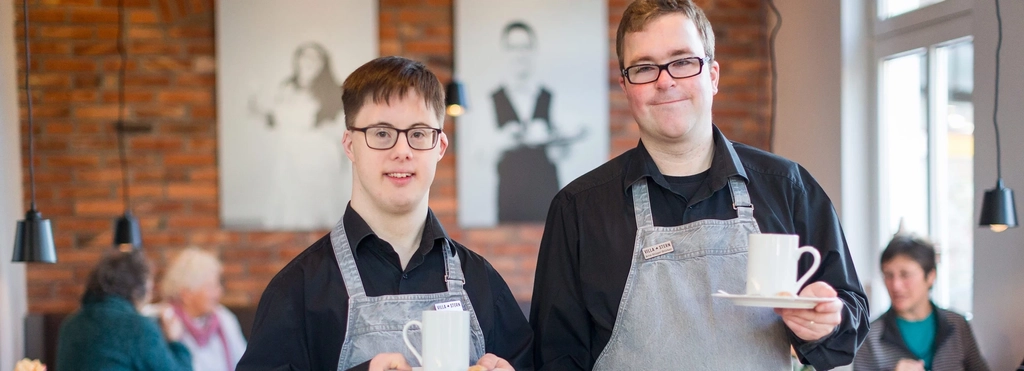 Zwei Männer mit Behinderung lächeln in die Kamera. Sie tragen Schürzen und halten Kaffeetassen in der Hand.