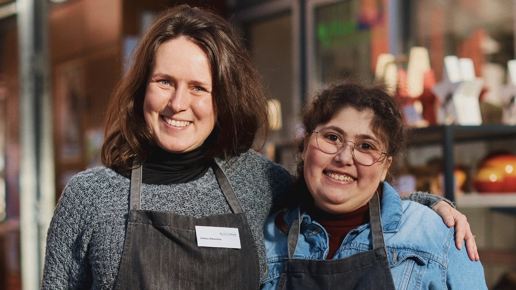 Zwei Frauen mit dunklen Schürzen stehen vor einem Marktstand. Eine hat den Arm freundschaftlich um die Schulter der anderen gelegt und beide lächeln in die Kamera.