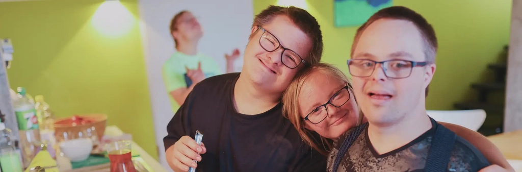 Drei junge Personen mit Down-Syndrom stehen Arm in Arm zusammen in der Küche am Herd und lächeln in die Kamera.